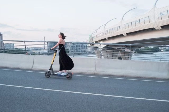 Woman in a black dress riding an electric scooter along an urban bridge and elevated highway.